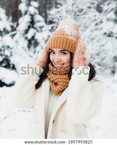 A beautiful happy laughing young girl in gloves and a winter hat-scarf, covered with snow flakes. Winter forest landscape background