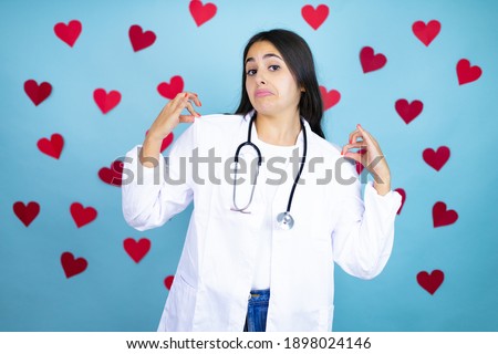 Young doctor woman wearing medical coat and stethoscope over blue background with red hearts holding her t-shirt with a successful expression