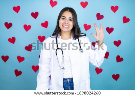 Young doctor woman wearing medical coat and stethoscope over blue background with red hearts doing hand symbol