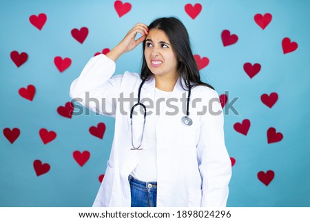 Young doctor woman wearing medical coat and stethoscope over blue background with red hearts confuse and wonder about question. Uncertain with doubt, thinking with hand on head