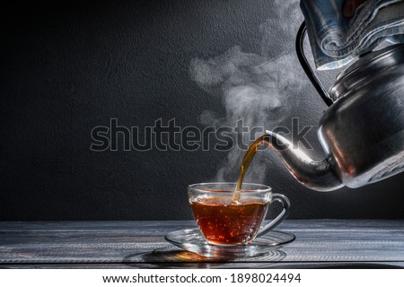 Pour the hot tea into the teacup. A teacup placed on an old wooden table In a black background, there was soft sunlight shining into a warm atmosphere. Royalty-Free Stock Photo #1898024494