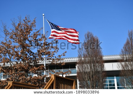 An American flag waving in the wind. A building is behind it. It is winter and the trees’ leaves are brown. Picture taken in O’Fallon, Missouri.