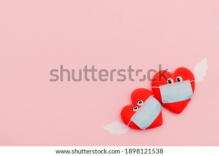 Two hearts with eyes and angel wings in protective masks on a pink background. Valentine's Day background. Coronavirus pandemic concept.Flat lay, top view,copy space.