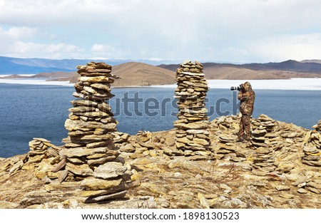 Lake Baikal in spring. Ice drift in the Small Sea Strait. Tourist in protective warm clothing takes pictures of tall stone pyramids on top of cliff against blue sky. Active outdoor and travel concept 