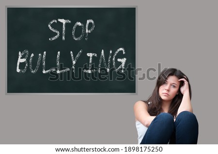 Sad teenage girl in front of a black chalkboard, stop bullying is written on the board, isolated in front of gray background