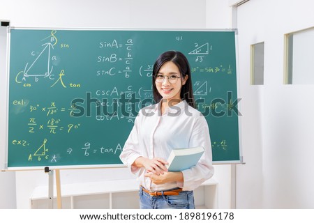 asian senior high school female math teacher hold books and smile at you in front of blackboard at classroom Royalty-Free Stock Photo #1898196178