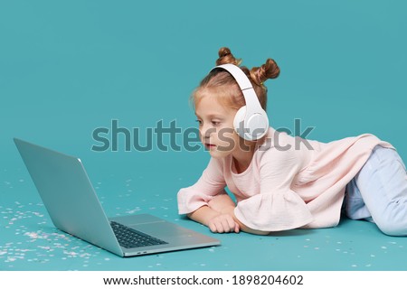 Smart child girl in wireless headphones using laptop on cyan background. Education, online study, tv, distance learning. Primary school student during quarantine. Educational games for children 