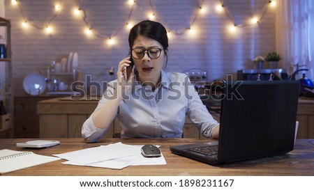 Angry female manager arguing and yelling at mobile phone while working on deadline project in evening at home.