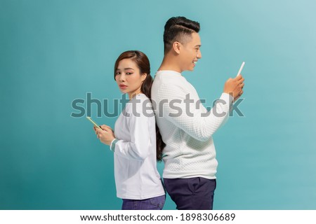 Asian girl and smile guy standing back to back, using mobile phones over blue background