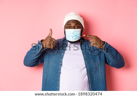 Coronavirus, lifestyle and global pandemic concept. Young african-american man pointing at face mask and showing thumbs-up, protect himself from covid, pink background