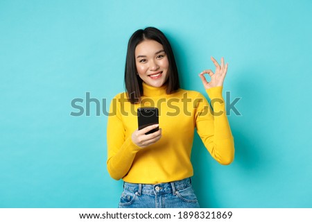 E-commerce and online shopping concept. Portrait of asian woman showing OK sign and using mobile phone, praise app, standing over blue background