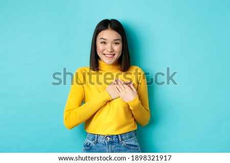 Image of beautiful asian woman holding hands on heart and smiling, thanking you, feeling grateful, standing over blue background