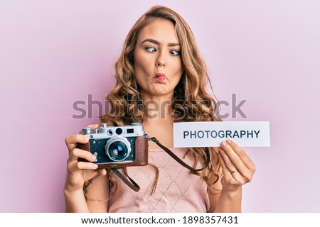 Young blonde girl holding vintage camera and paper with photography word paper making fish face with mouth and squinting eyes, crazy and comical. 
