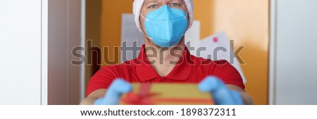 Male courier wearing medical protective mask and santa claus hat holds out a handy box. Safe services and service for delivery of goods and mail in the coronavirus pandemic concept