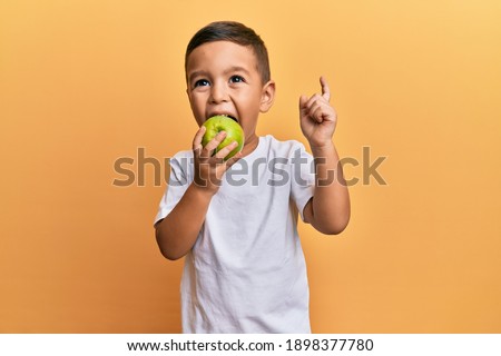 Adorable latin toddler smiling happy eating green apple looking to the camera over isolated yellow background. Royalty-Free Stock Photo #1898377780