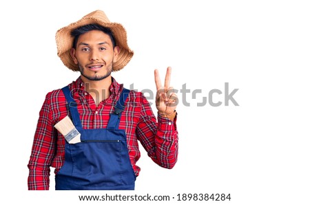 Handsome latin american young man weaing handyman uniform smiling looking to the camera showing fingers doing victory sign. number two. 