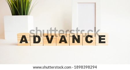 Wooden cubes with letters on a white table. The word is ADVANCE. White background with photo frame, house plant.