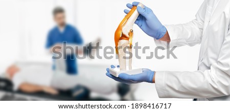 Physiotherapist holds an anatomical human knee-joint model in his hands, over background physiotherapy session for an elderly patient. Concept physio treatment and kinesiotherapy Royalty-Free Stock Photo #1898416717