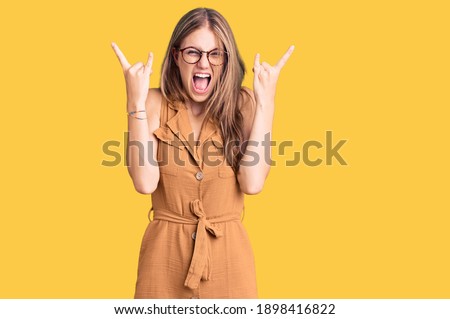Young beautiful blonde woman wearing casual clothes and glasses shouting with crazy expression doing rock symbol with hands up. music star. heavy music concept. 
