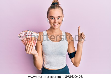 Beautiful blonde sport woman holding 100 norwegian krone banknotes smiling with an idea or question pointing finger with happy face, number one 