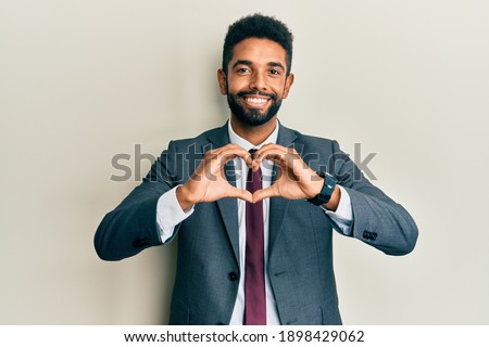 Handsome hispanic man with beard wearing business suit and tie smiling in love doing heart symbol shape with hands. romantic concept. 