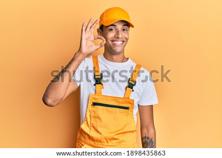 Young handsome african american man wearing handyman uniform over yellow background smiling positive doing ok sign with hand and fingers. successful expression. 