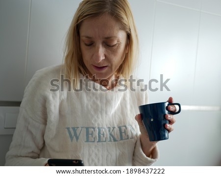 Shallow focus shot of a blonde woman using a smart phone and a cup in the kitchen with white wall background