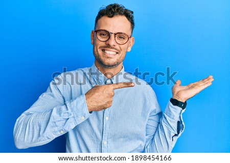 Young hispanic man wearing casual clothes and glasses amazed and smiling to the camera while presenting with hand and pointing with finger. 