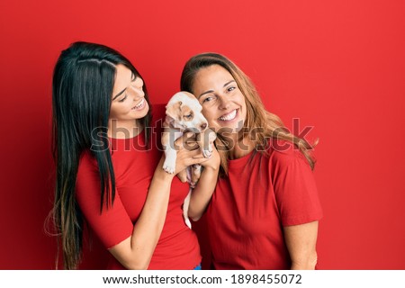 Beautiful hispanic mother and daughter smiling happy hugging little dog over isolated red background.
