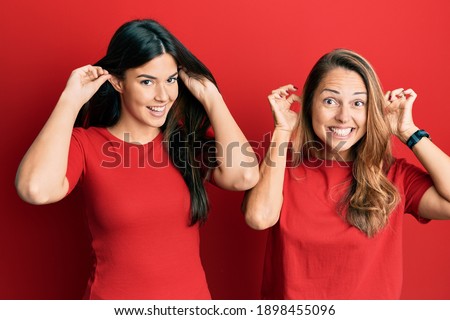 Hispanic family of mother and daughter wearing casual clothes over red background smiling pulling ears with fingers, funny gesture. audition problem 