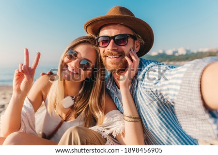 young attractive smiling happy man and woman in sunglasses sitting on sand beach taking selfie photo on phone camera, romantic couple by the sea on sunset, boho hipster style outfit