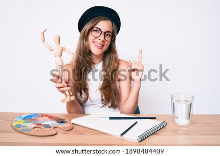 Beautiful young caucasian woman wearing artist look with beret holding manikin smiling happy pointing with hand and finger to the side 