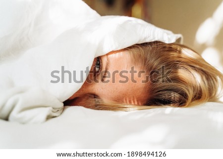 Beautiful, young blonde lies on the bed in the rays of soft, morning sun. Buried under the covers, showing out small part of her face. Wakes up slowly. Difficulty getting up, let me sleep some more. Royalty-Free Stock Photo #1898494126