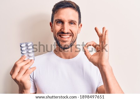 Young handsome man holding medicine pills standing over isolated white background doing ok sign with fingers, smiling friendly gesturing excellent symbol