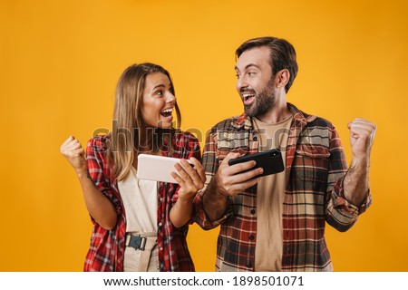 Excited couple making winner gesture while playing video game on cellphones isolated over yellow background