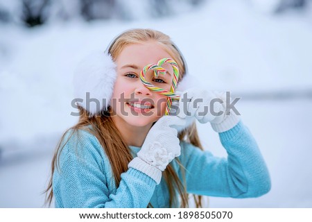 A girl in skates sits on a skating rink on the ice. In her hands are candy canes. Skating rink on the background of the winter forest. Selective focus photo