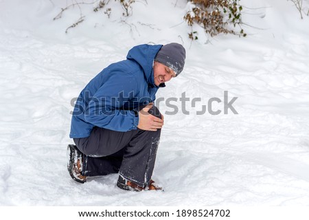Photo of injured young man bend down on snow and holding his knee in pain, outdoor. Young man holding his knee in pain on a snowy cold winter day. Man having a knee injury on winter road. Copy space.