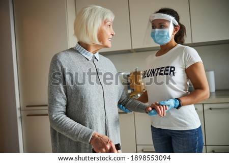 Dark-haired young carer in a face shield supporting a smiling senior woman by the arm