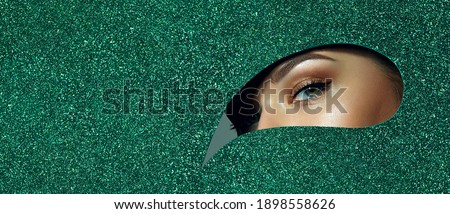 A girl with beautiful green eyes, with bright shadows on her eyelids. Looks through a teardrop hole made of green shiny paper.
