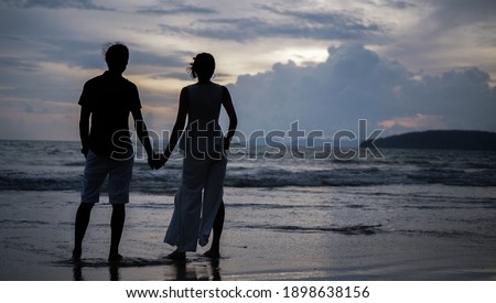 Romance near the ocean as a loving couple hold hands while looking out into the calm open sea. A silhouette photo of a lover holding hands at the beach with the ocean and cloudy sky in the background