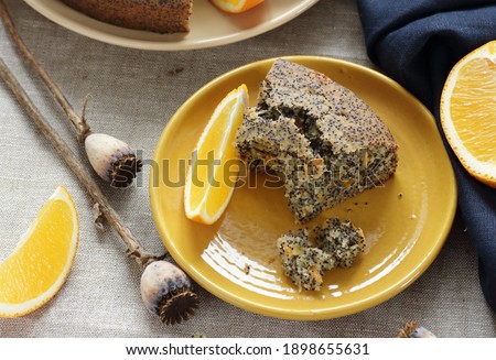 Poppy seed and orange peel vegan egg free and dairy free cake, bite on the orange plate with blue linen textile and poppy head nearby, closeup, copy space, vegan food and pastry concept