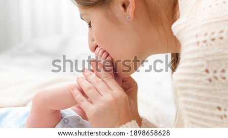 Closeup of loving and caring mother holding and kissing little feet of her newborn baby boy on bed. Concept of family happiness and loving parents with little children.