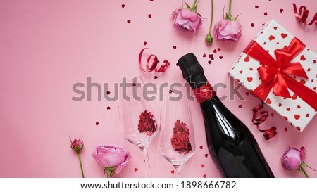 Concept for banner,greeting card for Valentine's day.Bottle of champagne wine,glasses with glittering confetti in shape of heart,roses,serpentine and gift box on pink background, flat lay,copy space