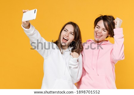 Joyful two young women friends 20s wearing casual white pink hoodies standing doing selfie shot on mobile phone clenching fists like winner isolated on bright yellow color background studio portrait