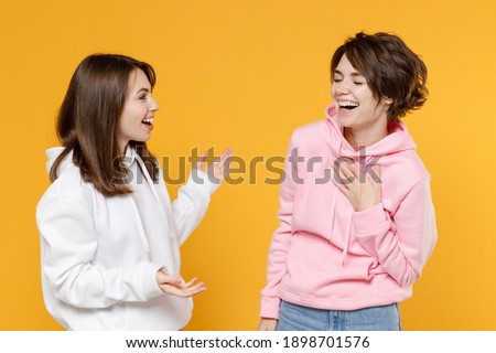 Laughing funny two young women friends 20s wearing casual white pink hoodies standing speaking talking spreading hands looking at each other isolated on bright yellow color background studio portrait Royalty-Free Stock Photo #1898701576