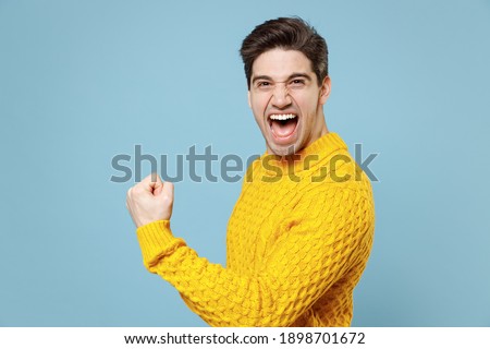 Young caucasian attractive gladden excited student man 20s in casual knitted yellow sweater doing winner gesture clench fists saying yes scream celebrating isolated on blue background studio portrait Royalty-Free Stock Photo #1898701672