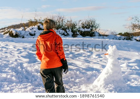 back view of a woman walking in the snow towards a snowman with a carrot in his nose on a mountain with a beautiful landscape and wearing an orange jacket making a very colorful image