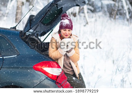 A cozy woman, a female in warm winter clothes, drinks a hot drink, tea or coffee, sits in the trunk of a car and smiles. Vacation, travel by car, snowy cold.