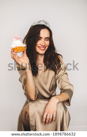 Image of brunette excited woman in tiara holding cakes while laughing isolated over white wall. Attractive woman wears beige dress and silver tiara, she hold cupcake with cream. Woman makes grimaces.