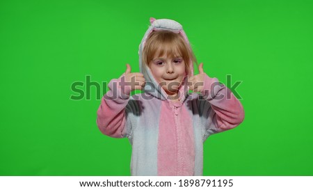 Young little blonde child 5-6 years old smiling, showing thumbs up gesture, agree sign in unicorn costume on chroma key green background. Portrait of kid girl animator in unicorn pajamas. Copy space